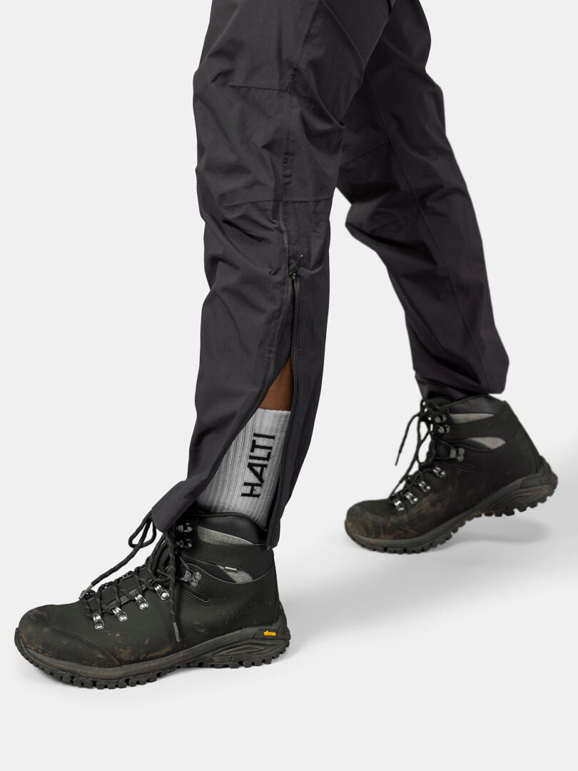 Person trägt schwarze Wanderschuhe, graue Halti-Socken und Halti Pisarat 2.5L DX Regenhosen Damen mit Knöchelreißverschlüssen.