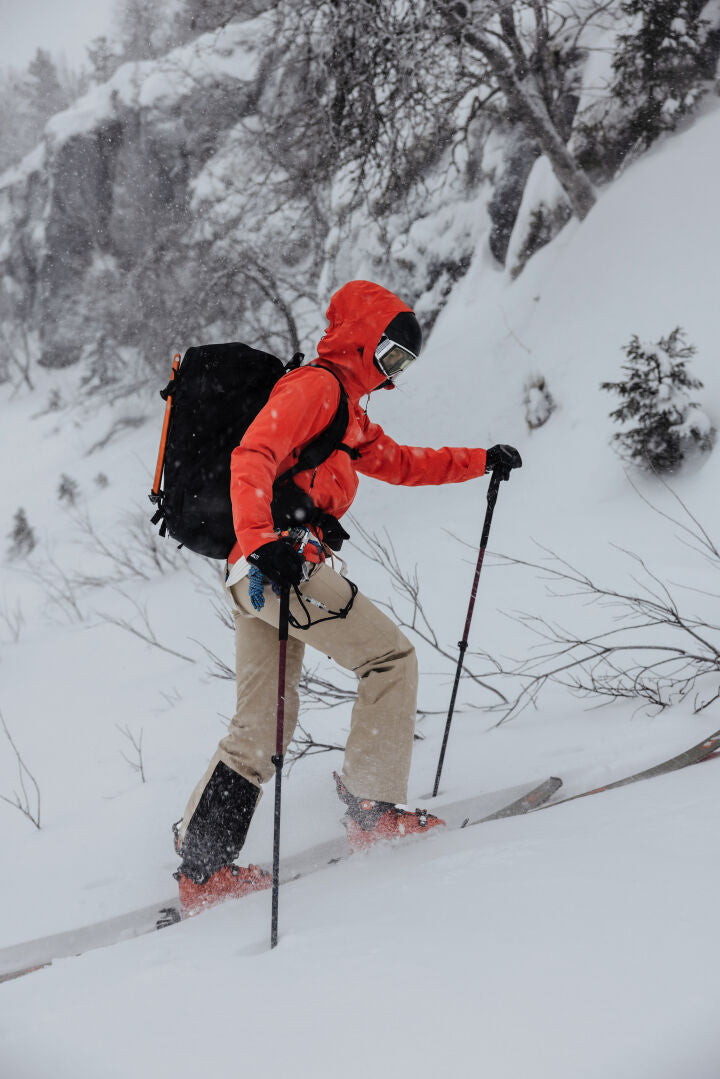Eine Person in roter Jacke, Halti Carvey II DX Skihose Damen und Skibrille fährt mit Stöcken und einem Rucksack bergauf durch den Schnee.