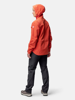 Person im Profil, die eine Halti Staala DrymaxX Regenjacke Damen in Orange, eine dunkle Hose und graue Schuhe vor einem einfarbigen Hintergrund trägt.
