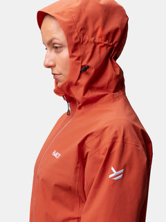 Eine Frau im Profil trägt die orangefarbene Halti Staala DX Regenjacke Damen mit Logo und Reißverschluss, vor weißem Hintergrund.