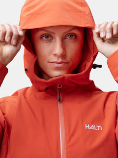 Eine Frau in einer orangefarbenen Halti Staala DrymaxX Regenjacke mit Kapuze hält die Seiten fest und zeigt das Halti-Logo.