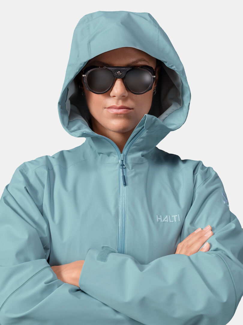 Eine Person in einer Halti Staala DX Regenjacke Damen, hellblau, verschränkt ihre Arme vor einem unifarbenen Hintergrund.