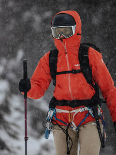 Eine Person in einer Halti Staala DrymaxX Regenjacke Damen und Skibrille steht mit einem Skistock und einer Kletterausrüstung im fallenden Schnee.
