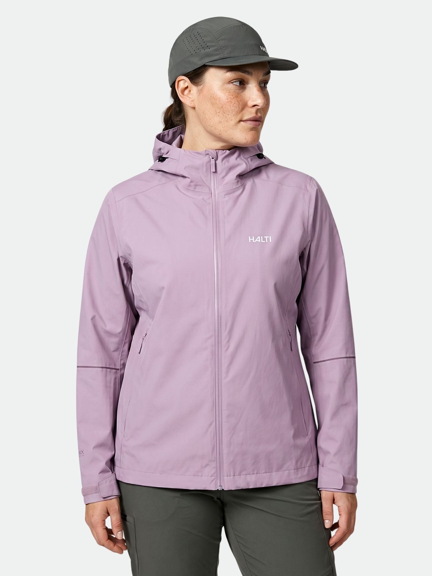 Eine Person in einer hellvioletten Halti Staala DX Regenjacke Damen Plus, einer grauen Hose und einer Mütze steht vor einem einfarbigen Hintergrund.