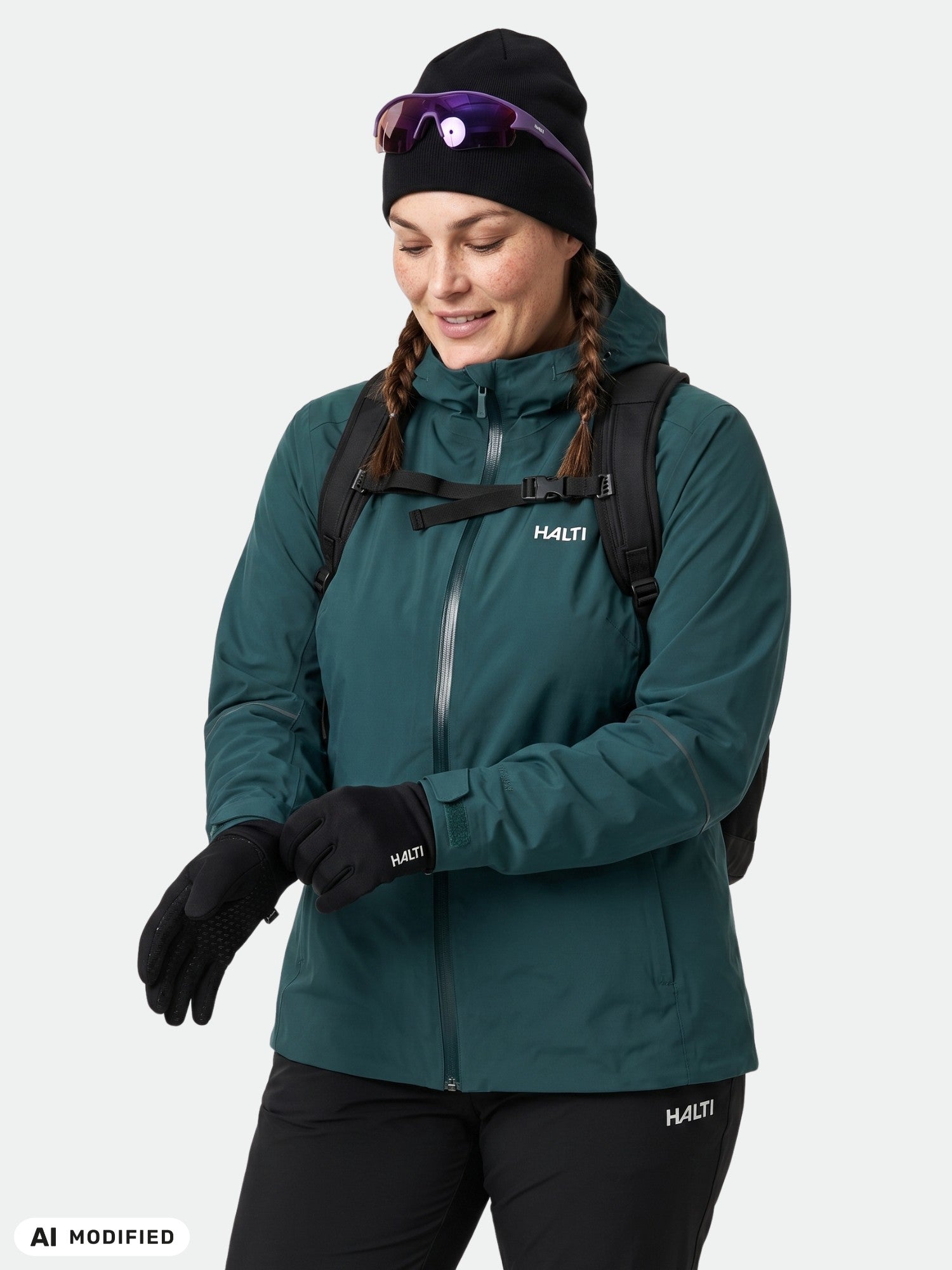 Eine Frau in Outdoor-Kleidung trägt eine grüne Halti Staala DX Regenjacke Damen Plus, Handschuhe, einen Rucksack und eine Sonnenbrille.