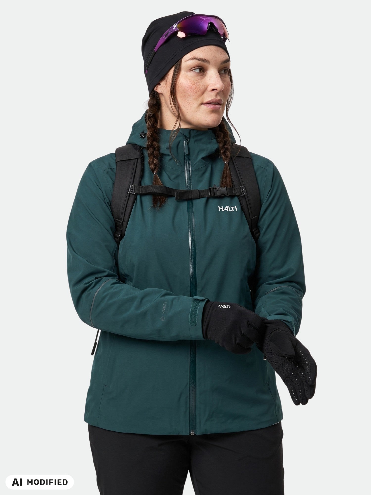 Frau in einer „Halti Staala DX Regenjacke Damen Plus“, schwarzer Mütze, Rucksack und Sonnenbrille auf dem Kopf, die zur Seite blickt.