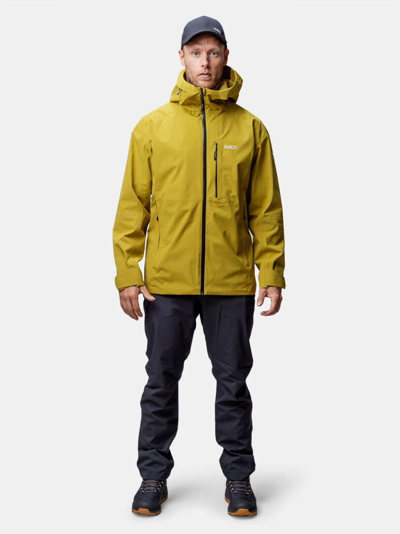 Ein Mann trägt eine gelbe Halti Staala DX Regenjacke Herren, eine schwarze Hose und eine dunkle Mütze vor einem schlichten Hintergrund.