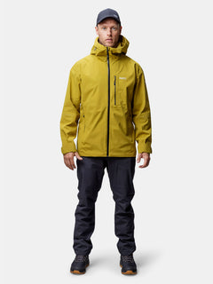 Ein Mann trägt eine gelbe Halti Staala DX Regenjacke Herren, eine schwarze Hose und eine dunkle Mütze vor einem schlichten Hintergrund.