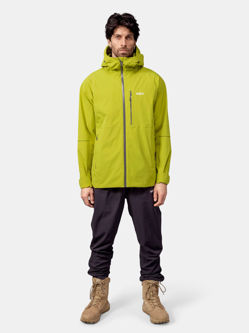 Ein Mann trägt eine lindgrüne Halti Staala DX Regenjacke Herren, eine schwarze Hose und braune Stiefel vor einem schlichten Hintergrund.