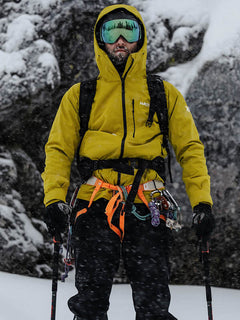 Eine Person in einer gelben Halti Staala DX Regenjacke Herren mit Kletterausrüstung steht im Schnee und hält Trekkingstöcke.