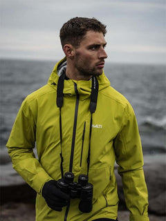 Ein Mann in einer Halti Staala DX Regenjacke Herren mit Fernglas steht am Meer, bereit für Abenteuer.