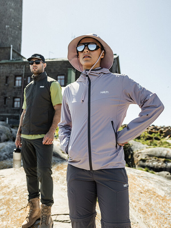 Zwei Personen in Halti Pallas Cool X-stretch Jacke Damen stehen auf einem Felsen vor einem Steingebäude, bereit für den Spaß im Freien.