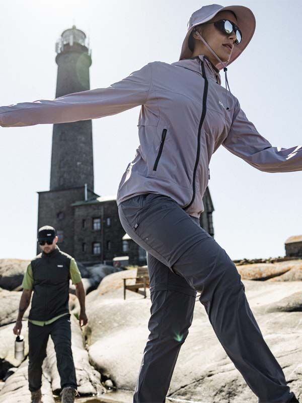 Zwei Personen in Halti Pallas Cool Stretch Zip-off Hosen Damen spazieren an einem sonnigen Tag über Felsen in der Nähe eines hohen Leuchtturms.