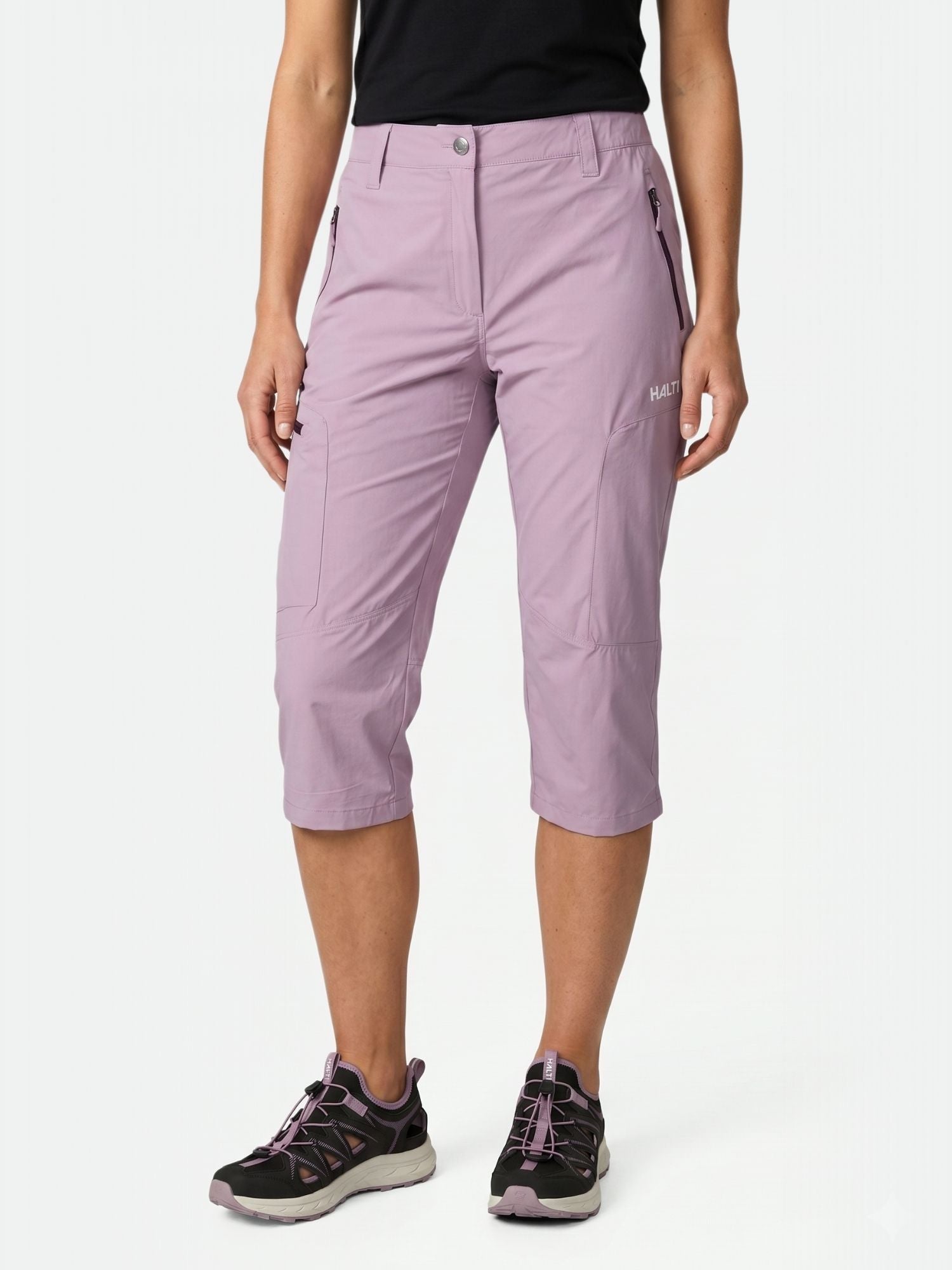 Eine Person in Halti Pallas Cool X-stretch Caprihose Damen, schwarzem Top und Wanderschuhen steht vor einem weißen Hintergrund.