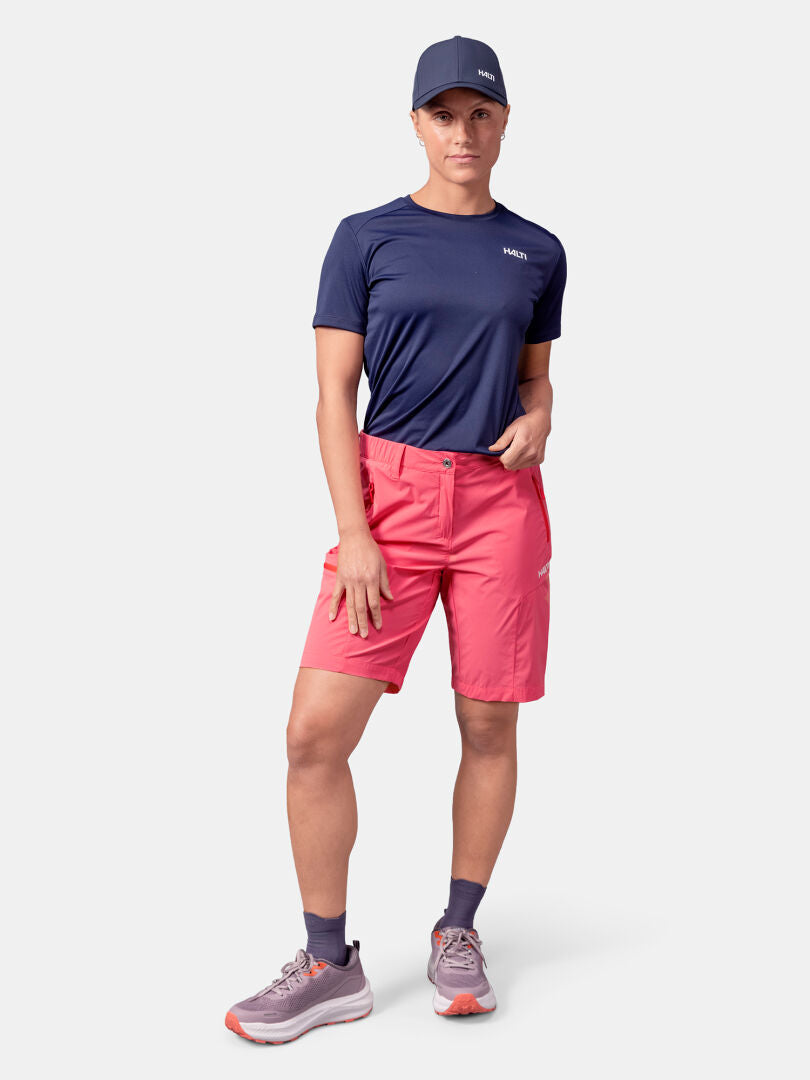 Person mit marineblauer Mütze, Halti Pallas Cool Shorts Damen und marineblauem T-Shirt steht vor einem einfarbigen Hintergrund.