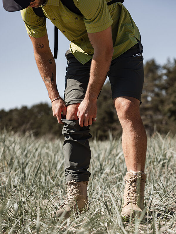 Ein Mann in Stiefeln und Shorts zieht die Halti Pallas Cool Stretch Zip-off Hosen Herren auf einer Wiese an.