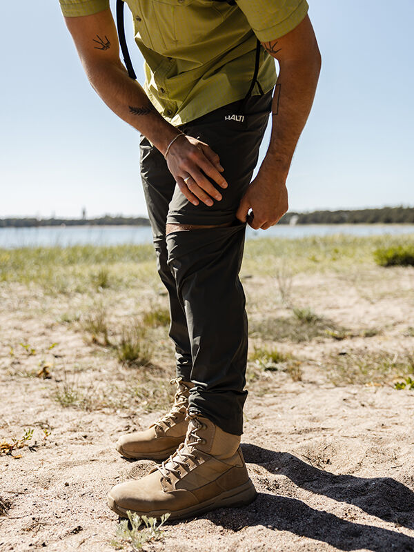 Draußen auf Sand, Person trägt Halti Pallas Cool Stretch Zip-off Hosen Herren und Wanderschuhe, Hände auf einem Bein.