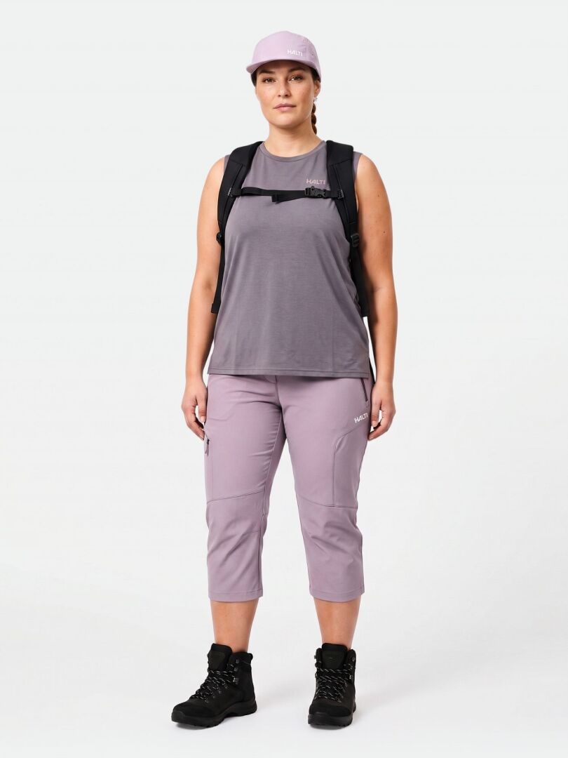 Eine Person steht mit Blick nach vorne in Halti Pallas Cool X-stretch Caprihose Damen Plus, einer lila Mütze und schwarzen Stiefeln.