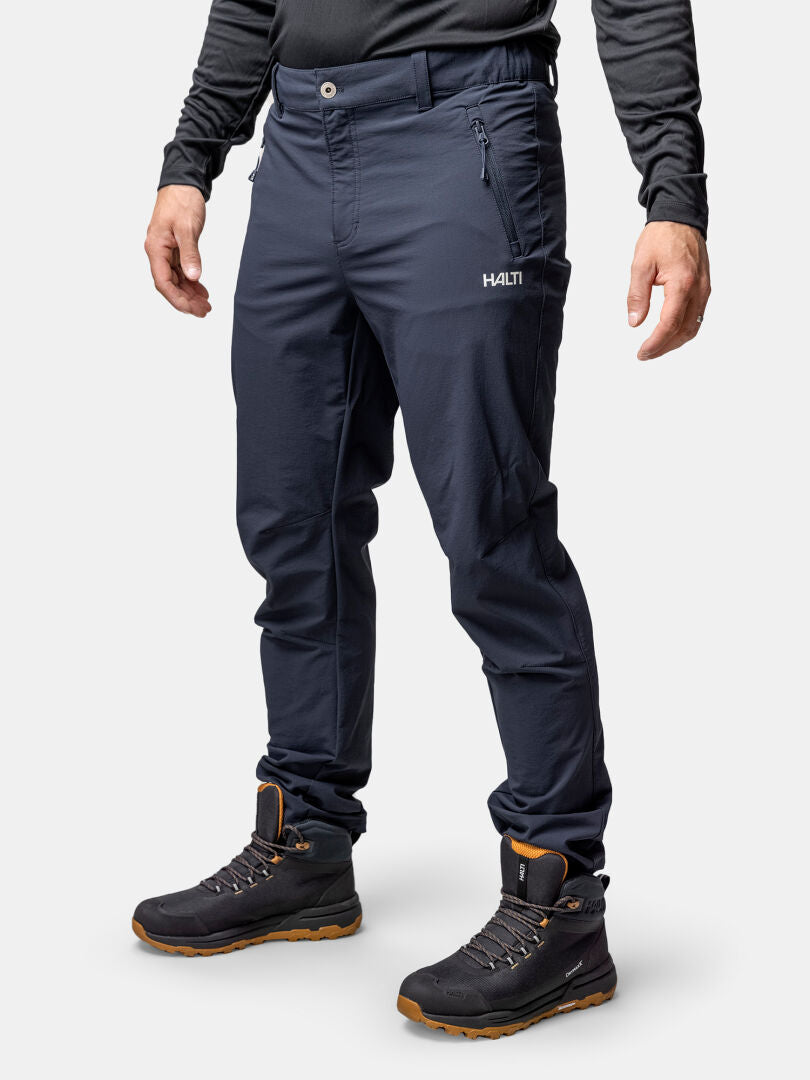 Eine Person trägt Halti Finholma X-stretch Hosen Herren, ein schwarzes langärmeliges Hemd und Wanderschuhe vor einem schlichten Hintergrund.