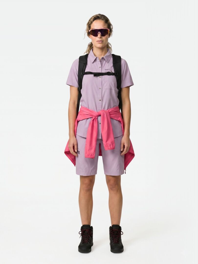 Eine Frau in Halti Pallas Cool Shorts steht mit dem Gesicht nach vorne, rosa Jacke in der Taille, schwarze Stiefel, Rucksack, Sonnenbrille.