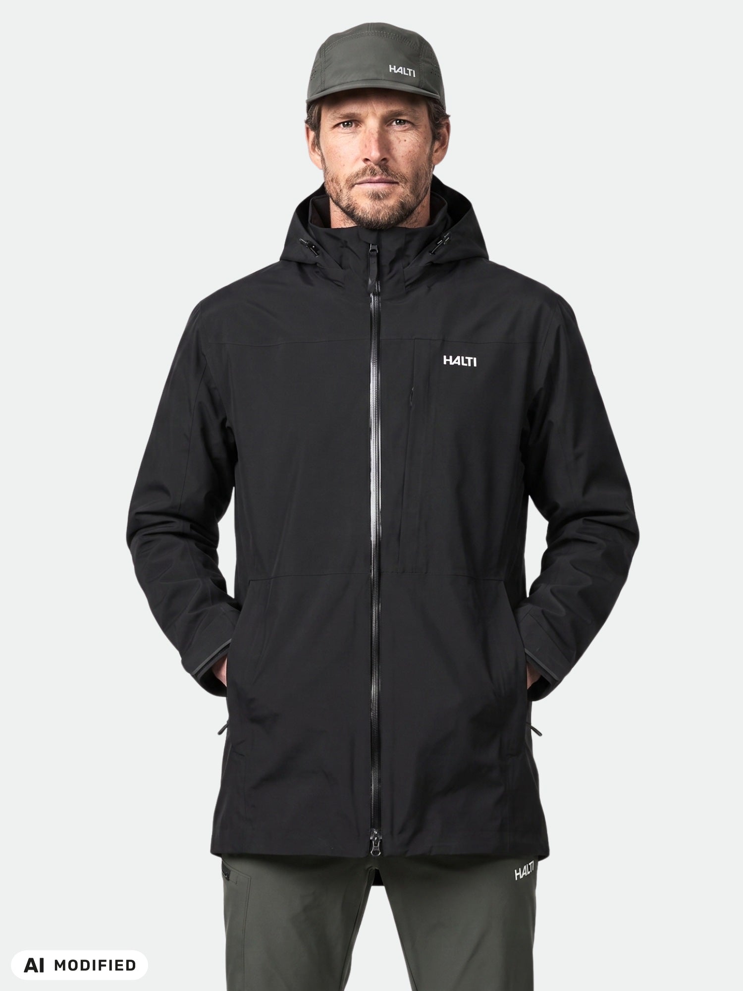 Ein Mann in einer schwarzen Halti Valaja DX Parkajacke und einer grünen Halti-Mütze, der vor einem einfarbigen, hellen Hintergrund steht.
