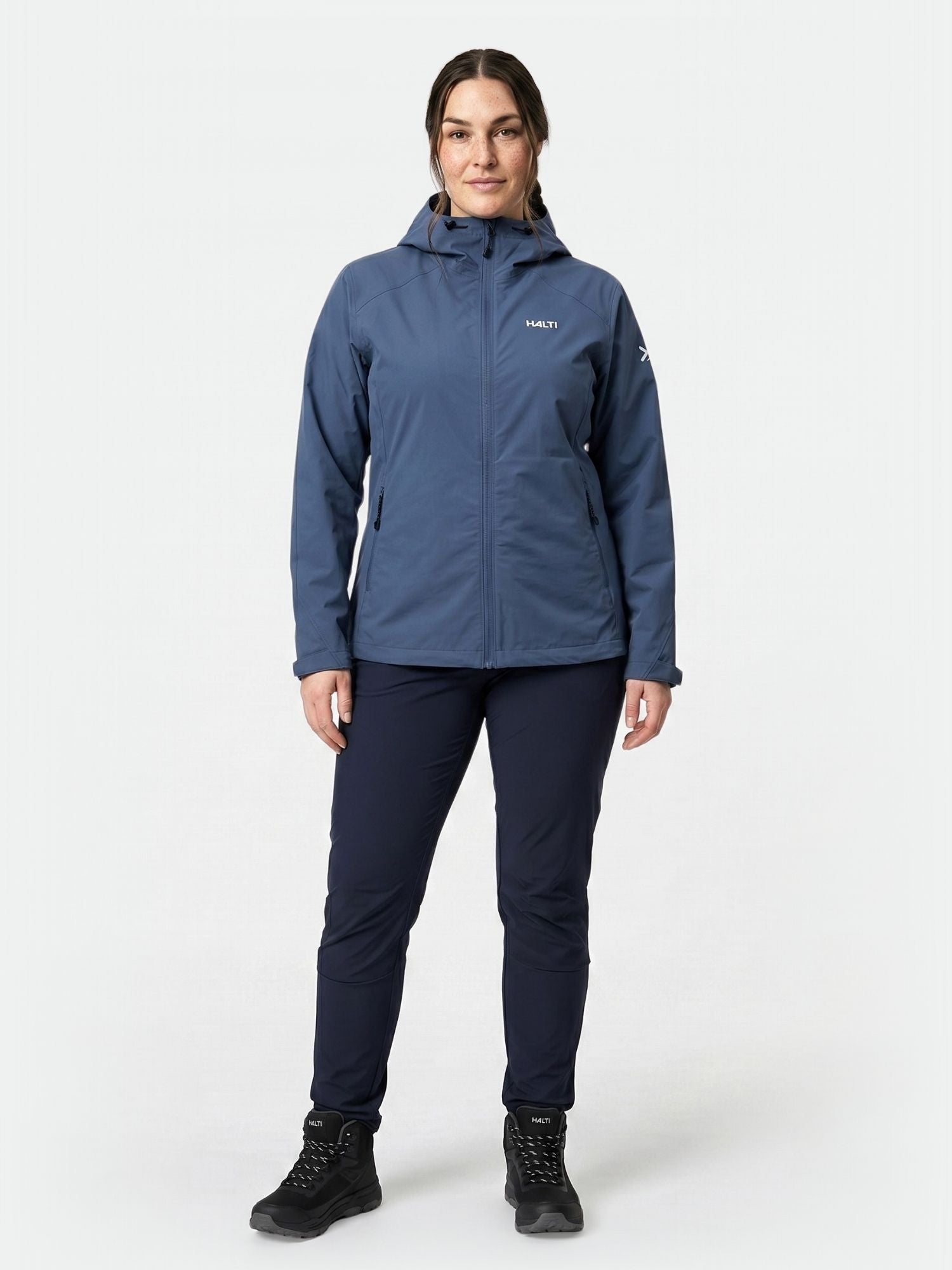 Eine Person trägt eine blaue Halti Finholma X-Stretch-Jacke für Damen in Größe Plus, eine marineblaue Hose und schwarze Stiefel vor einem schlichten, hellen Hintergrund.