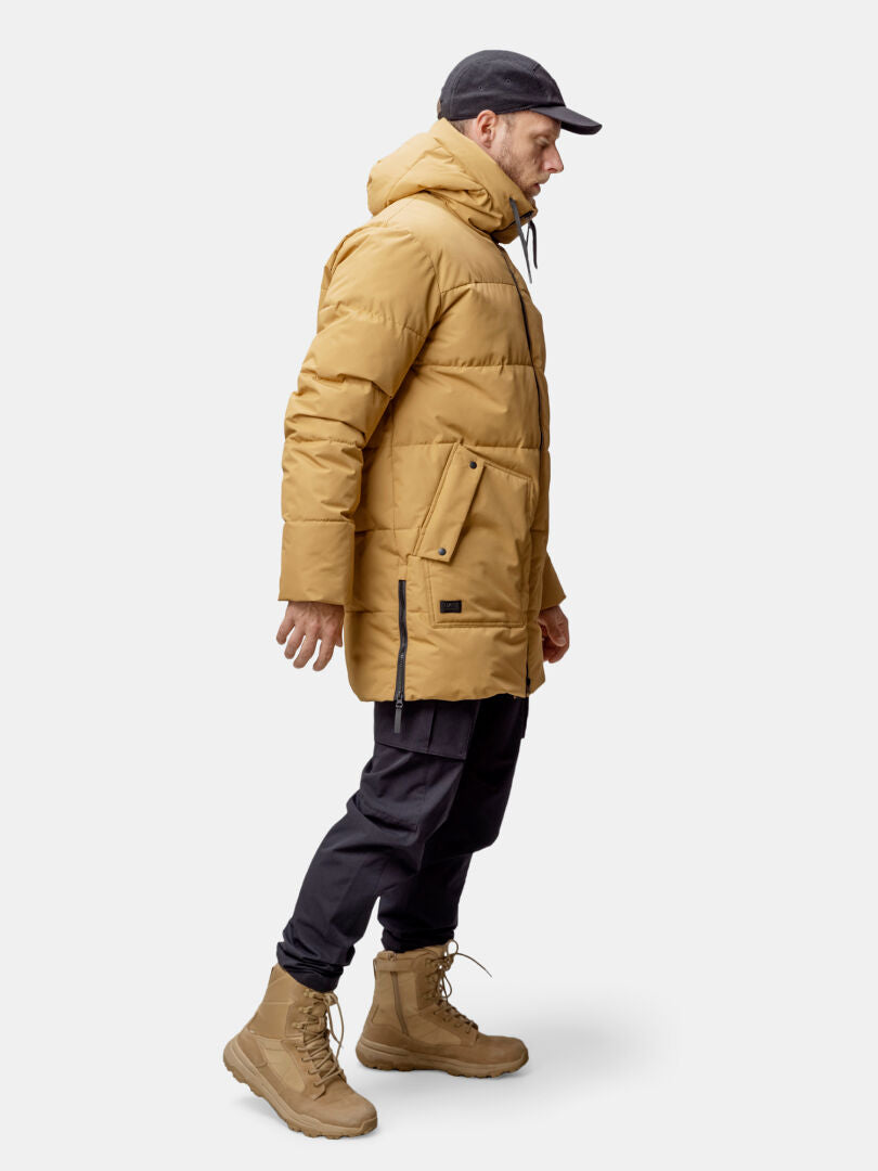Ein Mann in einer hellbraunen Halti Linjat Parka Jacke Herren mit Thermofill-Isolierung steht vor einem schlichten Hintergrund.