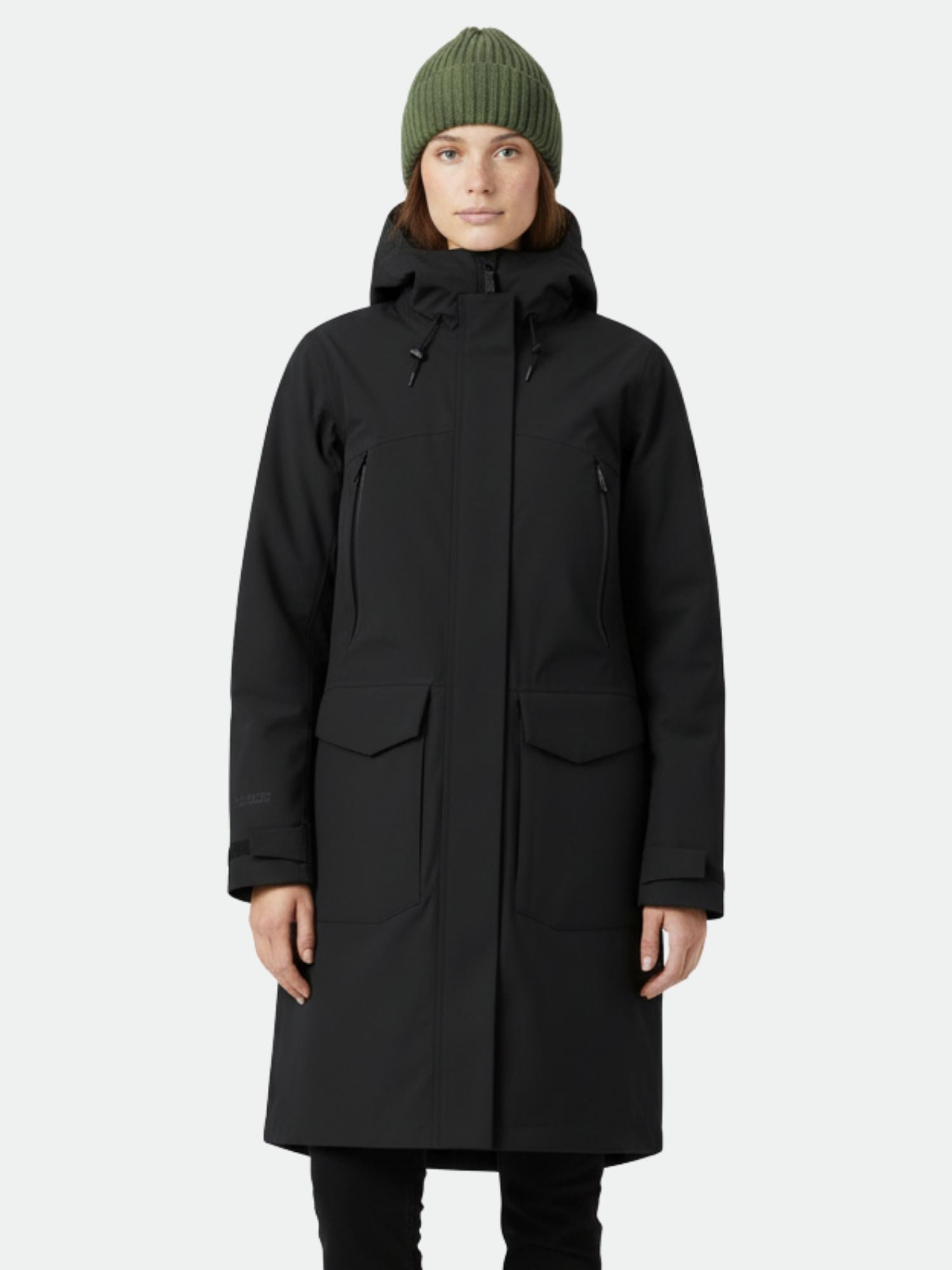 Eine Person im Halti Bergga DX Winter Parka Damen und einer grünen Strickmütze steht vor einem schlichten Hintergrund.