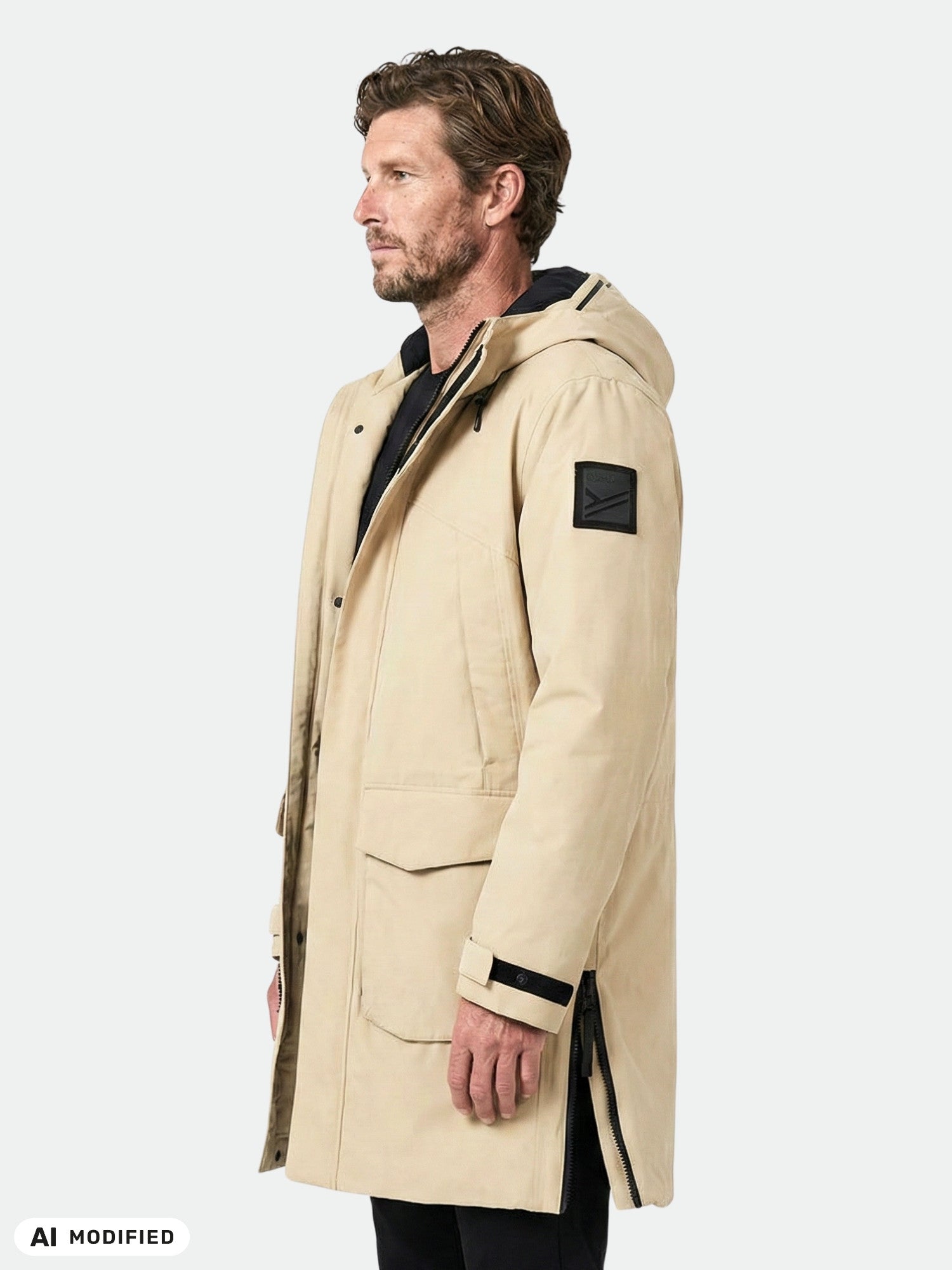 Ein Mann in einem beigen Halti Bergga DX Winterparka steht vor einem einfarbigen Hintergrund und blickt nach links, die Hände locker an den Seiten.