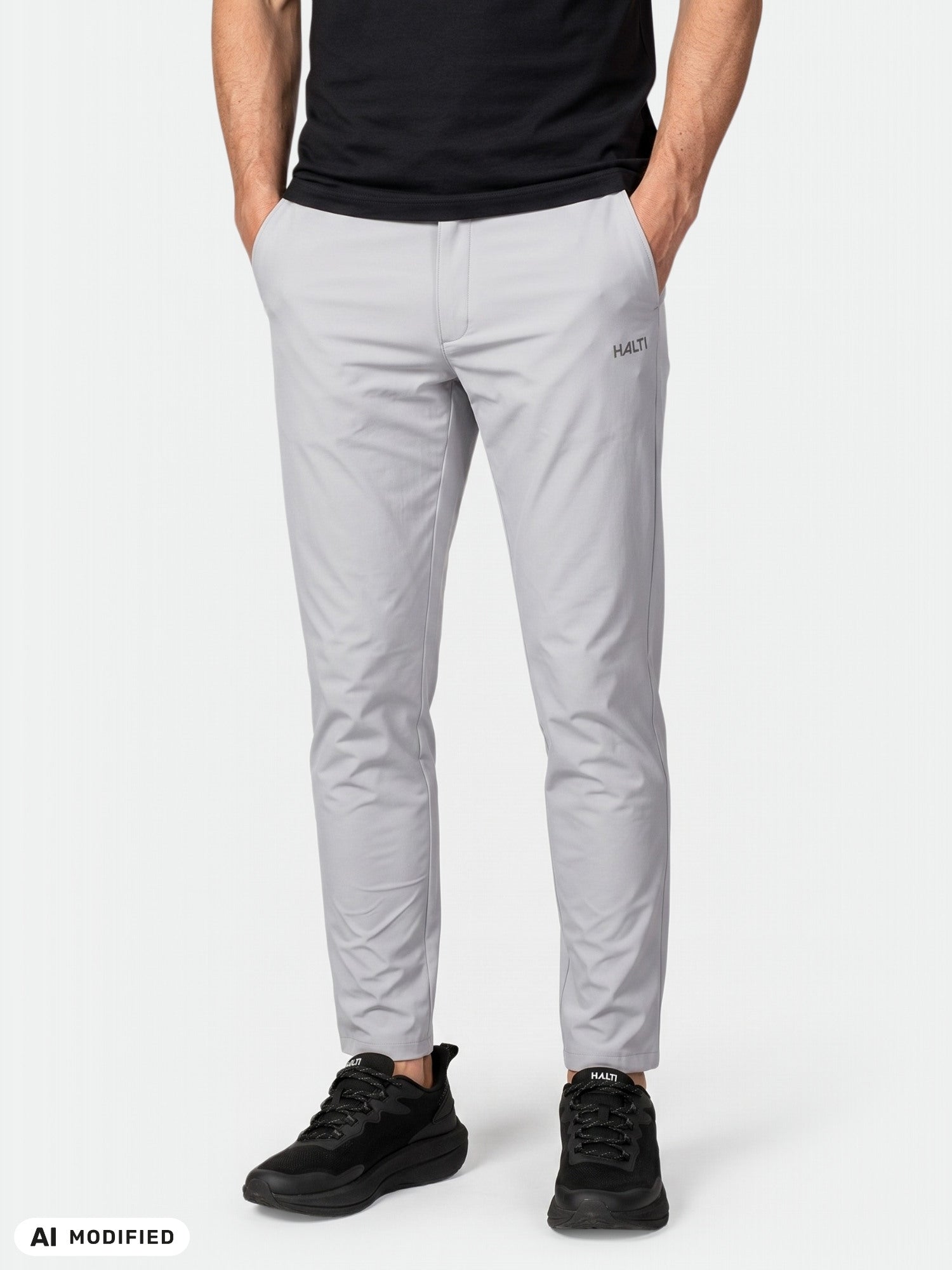 Ein Mann in hellgrauen Halti Drive X-Stretch-Chinos steht mit den Händen in den Taschen vor einem einfarbigen Hintergrund.
