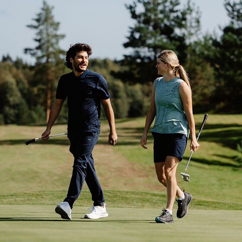 Ein Mann und eine Frau gehen auf einem Golfplatz in Halti Drive X-stretch Chinos Herren, jeder hält einen Schläger, dahinter Bäume.