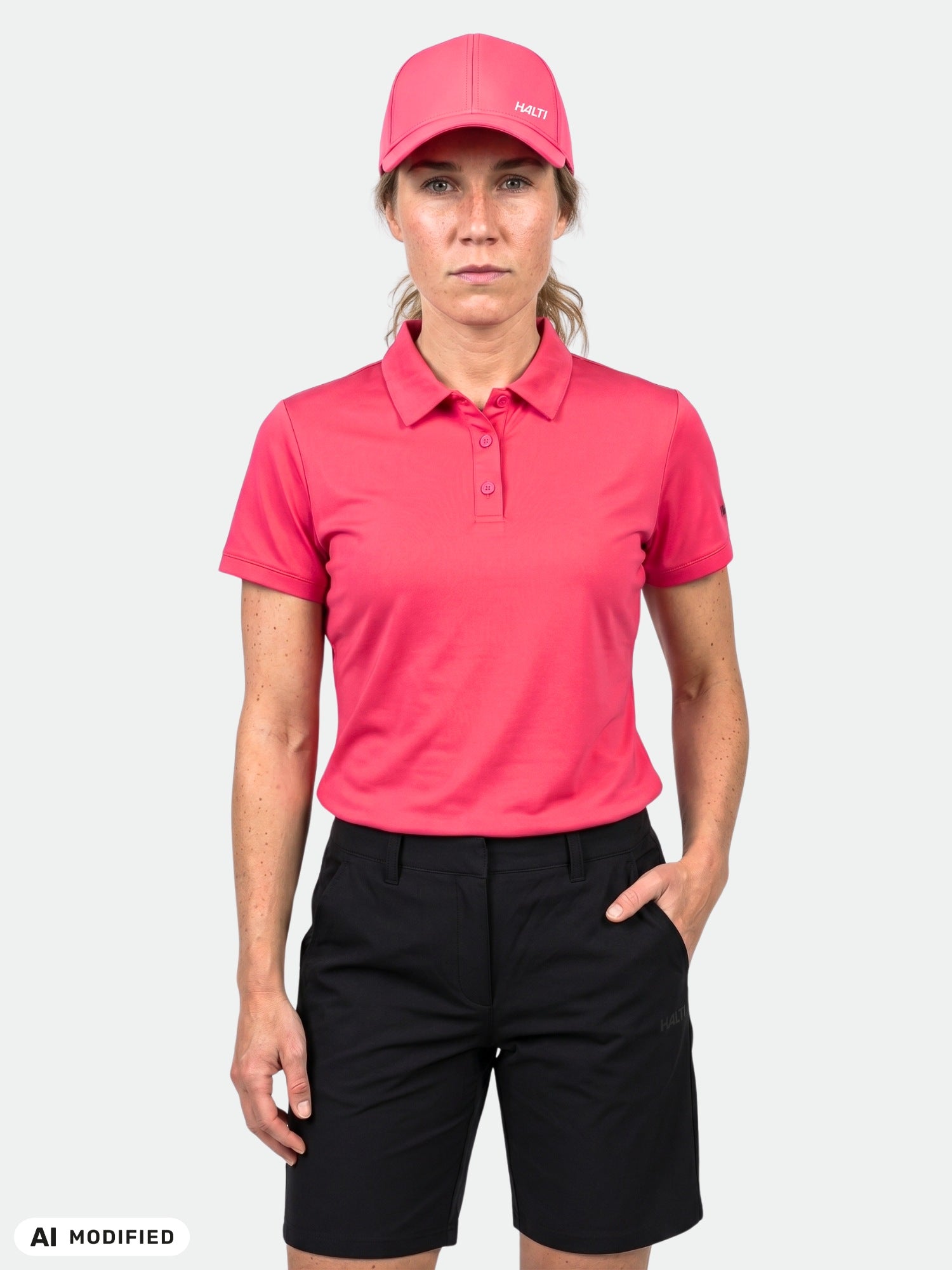 Ein Model in einem „Halti Laine Technisches Poloshirt“ für Damen, einer rosa Mütze und schwarzen Shorts steht mit dem Gesicht zur Kamera vor einem weißen Hintergrund.