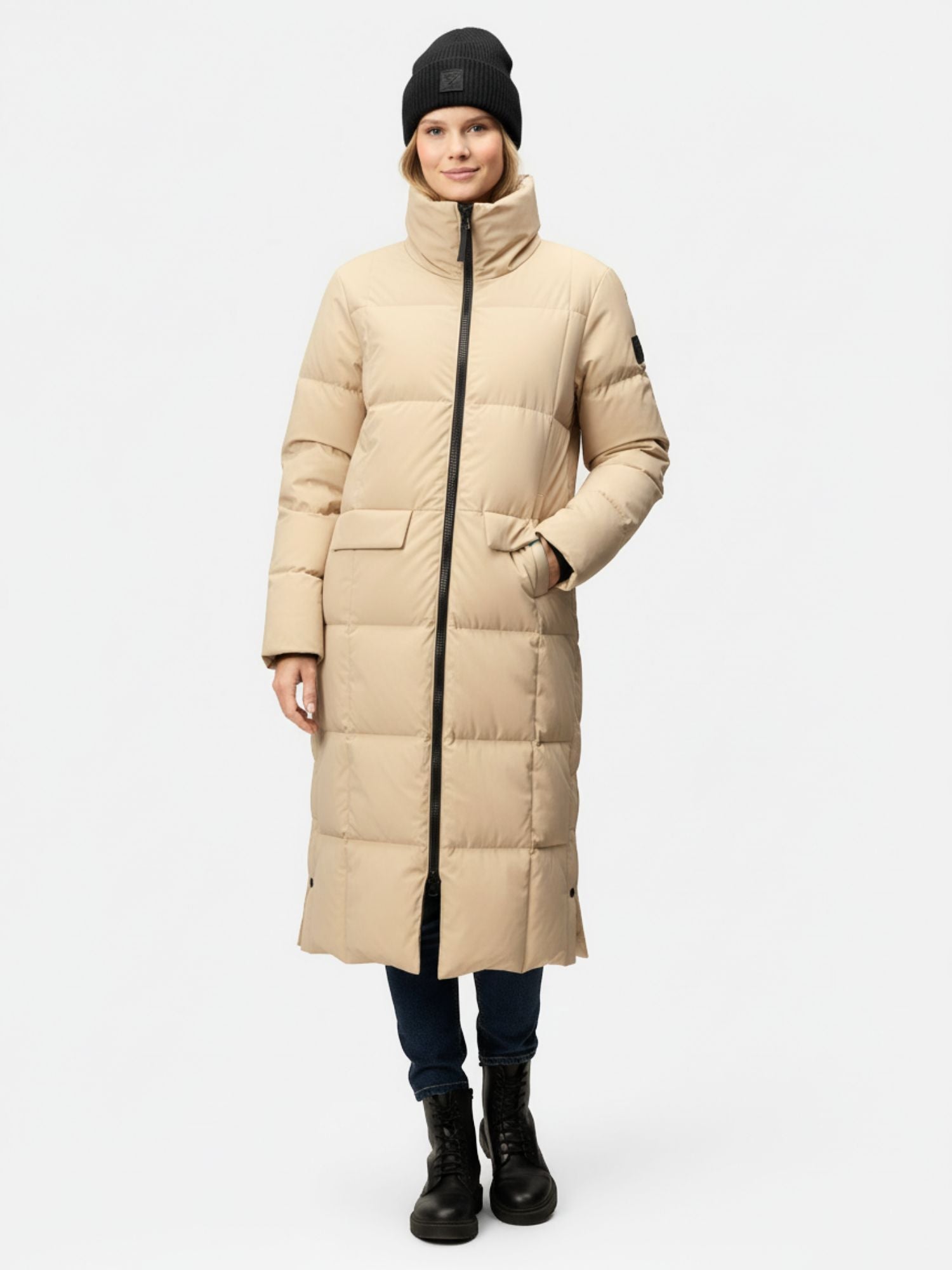 Eine Person trägt eine lange beige Halti Penger Winterjacke Damen, eine schwarze Mütze, dunkle Jeans und nach vorne gerichtete Stiefel.