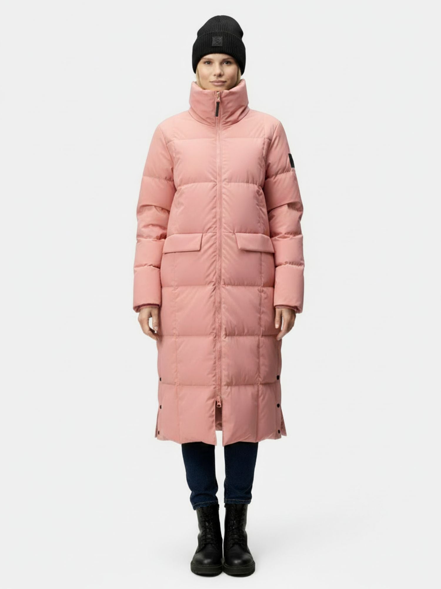 Eine Person in einer rosafarbenen Halti Penger Winterjacke Damen, einer dunklen Hose, schwarzen Stiefeln und einer schwarzen Mütze sieht nach vorne.