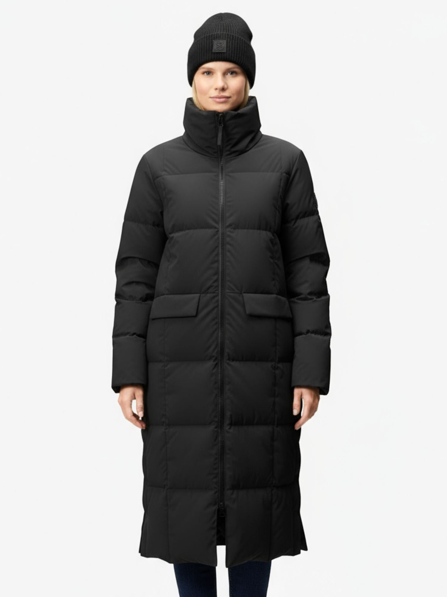 Eine Frau trägt eine lange schwarze Halti Penger Winterjacke Damen und eine schwarze Mütze vor einem schlichten weißen Hintergrund.