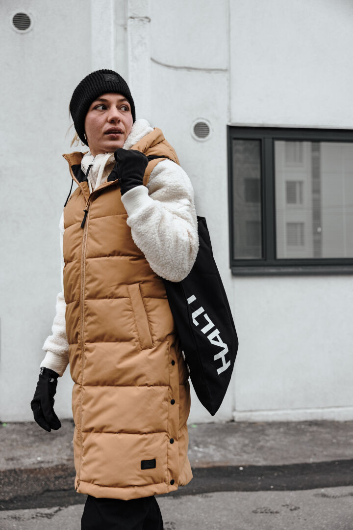 Person in hellbrauner Pufferweste und Halti Lykky Fleecepullover Damen mit einer schwarzen Tragetasche, vor einem grauen Gebäude.