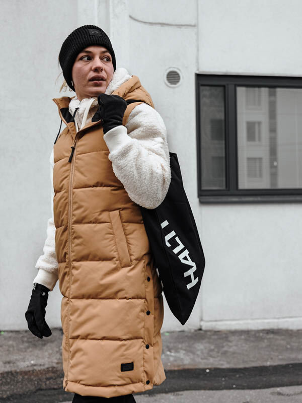 Die Person trägt eine hellbraune Halti Penger Puffer Weste Damen, einen weißen Kapuzenpulli, eine schwarze Mütze, Handschuhe und hält eine schwarze Halti-Tasche.