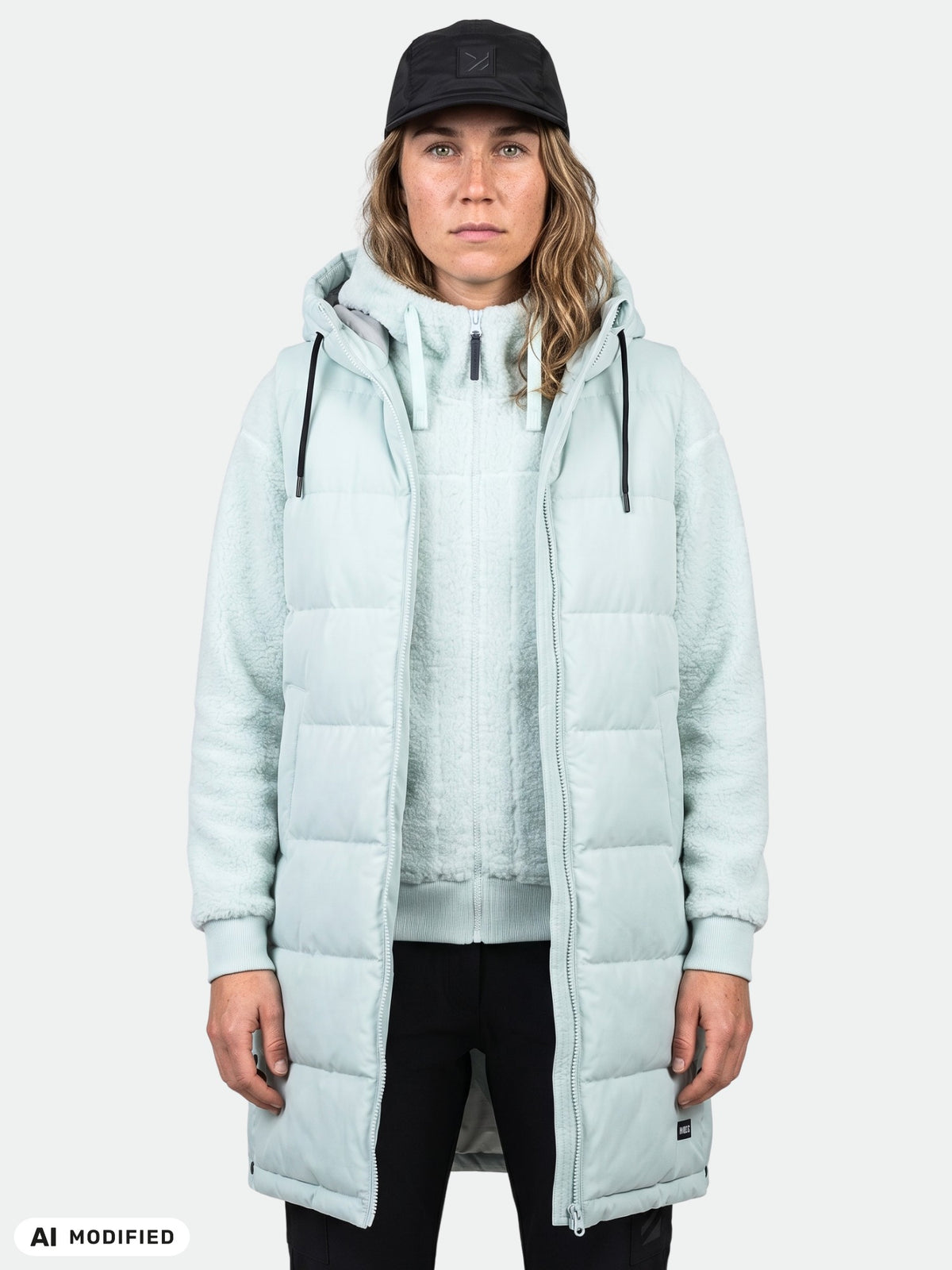 Eine Person trägt die „Halti Penger Puffer Weste Damen“ über einem Hoodie, dazu eine schwarze Hose und eine Mütze, vor einem einfarbigen Hintergrund.