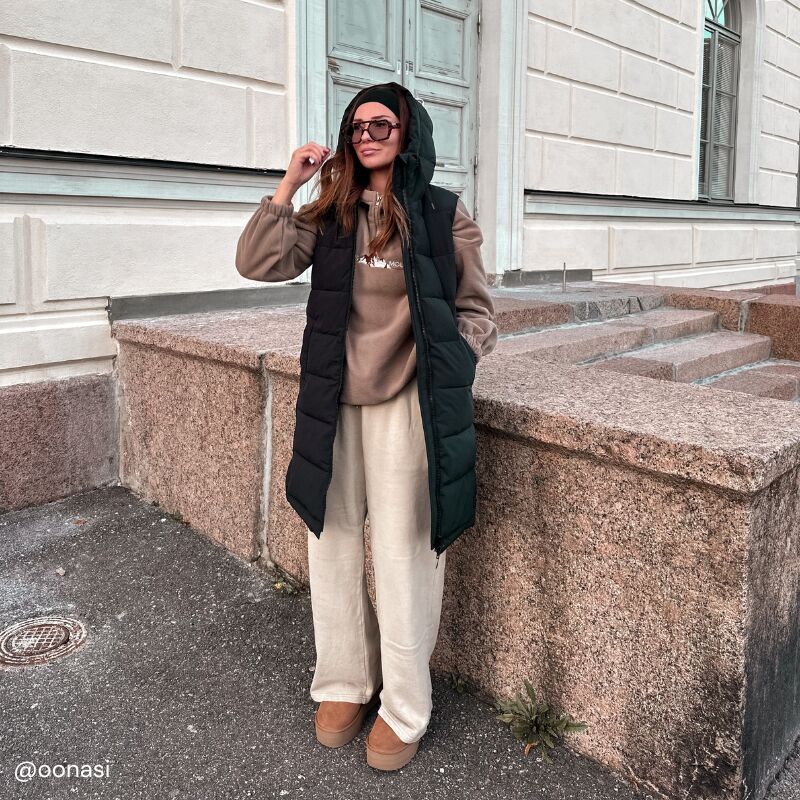 Eine Person steht draußen an einer Steintreppe und trägt die Halti Penger Puffer Weste Damen, ein braunes Sweatshirt, eine beige Hose und braune Schuhe.