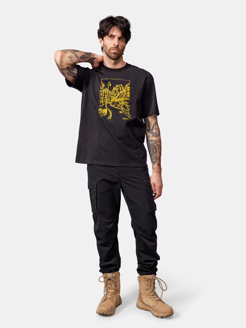 Mann in einem Halti Skaltsi Unisex T-Shirt, schwarzer Cargohose und hellbraunen Stiefeln, die Hand im Nacken.