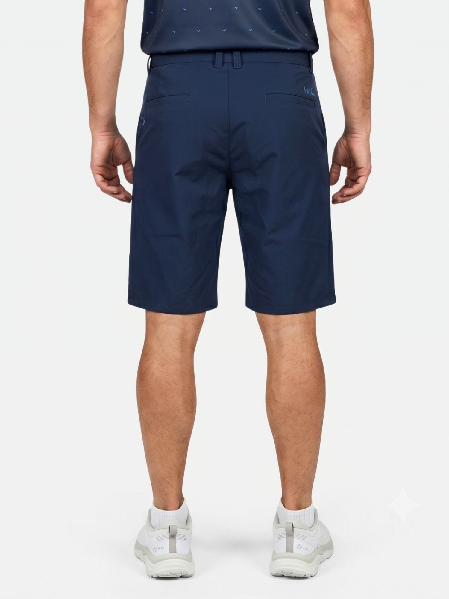 Ein Mann steht mit dem Rücken zur Kamera und trägt vor einem hellen Hintergrund die „Halti Drive Stretch Shorts Herren“, ein dunkles Hemd und weiße Sneaker.