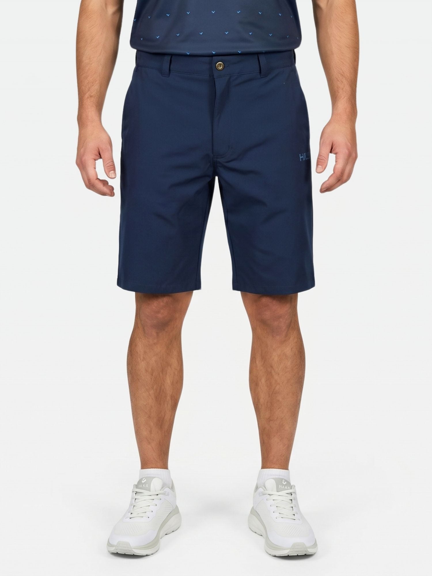 Eine Person in marineblauen „Halti Drive Stretch Shorts Herren“, einem dunklen Hemd und weißen Sneakers steht vor einem einfarbigen Hintergrund.