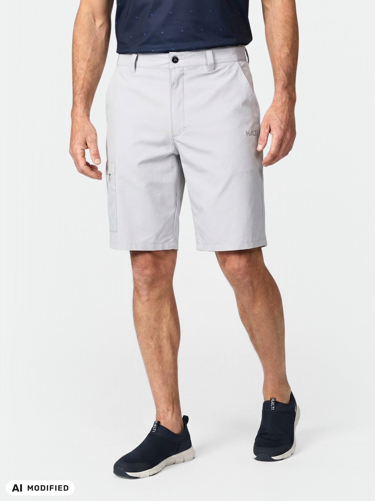 Ein Mann in „Halti Drive Stretch Shorts Herren“, einem marineblauen Hemd und schwarzen Slip-Ons steht vor einem einfarbigen Hintergrund.