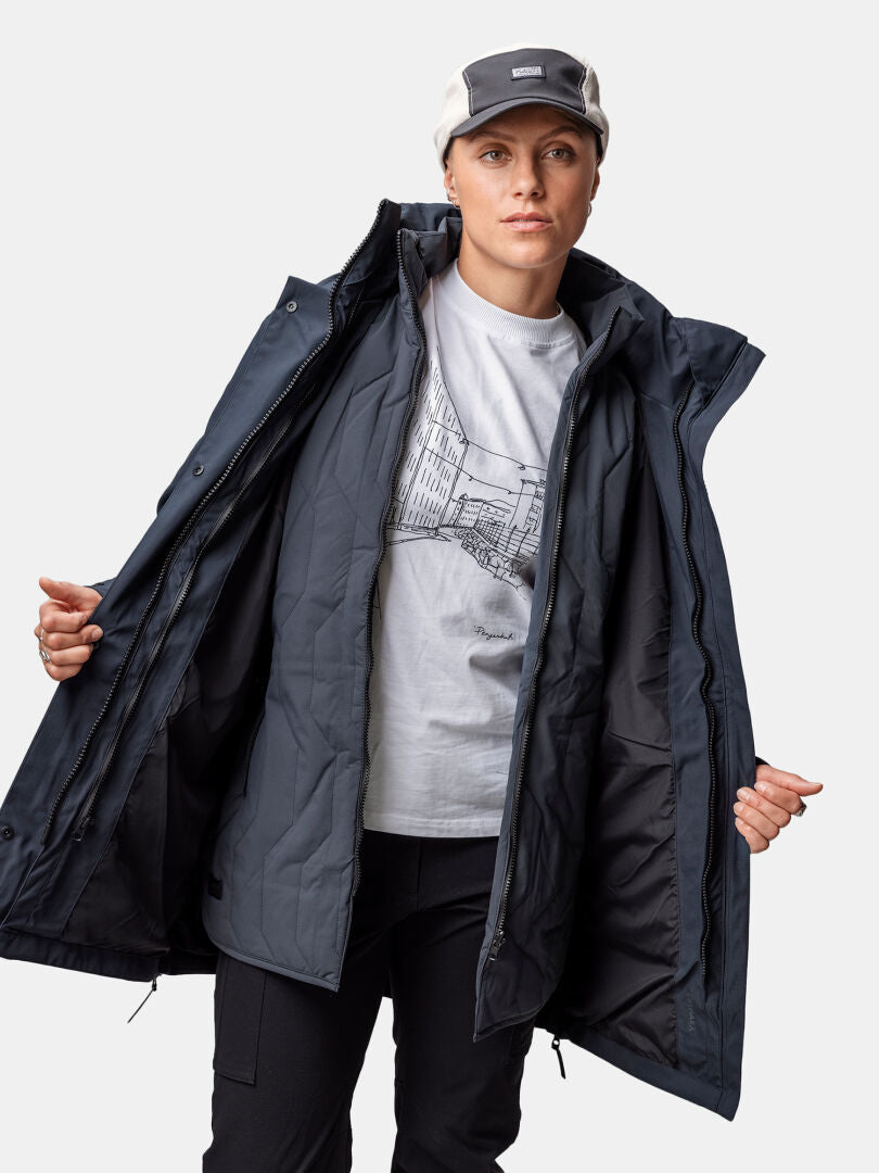 Outdoor-Enthusiastin mit grauer Cap, weißem Printshirt und offenem Halti Synkas 3in1 DrymaxX Parka Damen vor weißem Hintergrund.