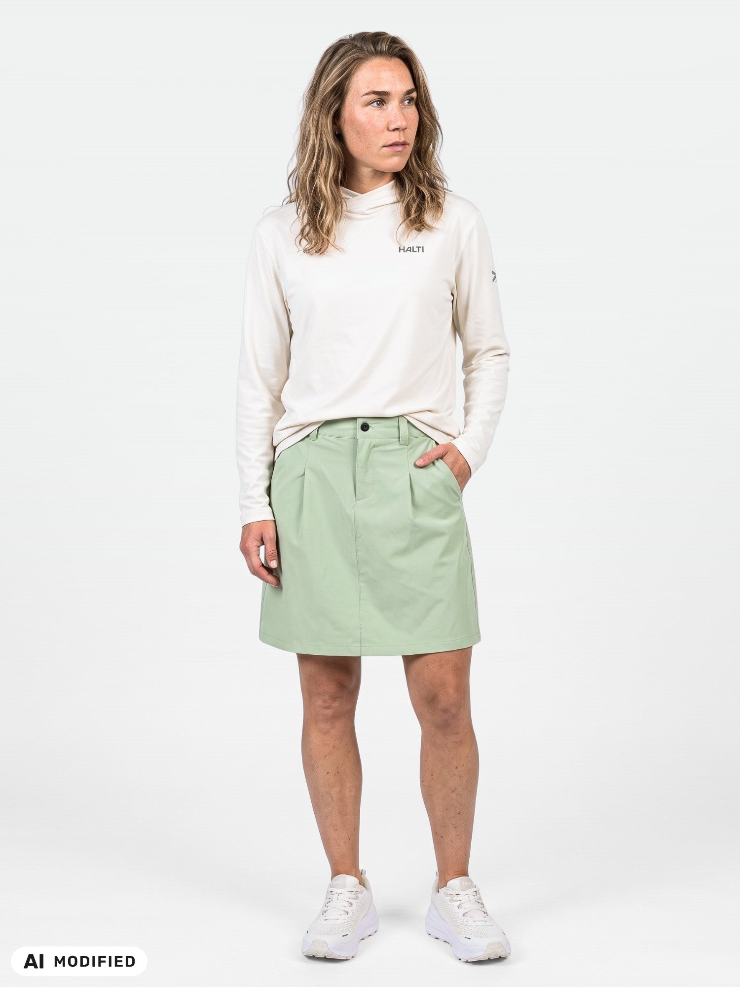 Eine Frau in einem weißen Langarmshirt und einem hellgrünen „Halti Drive X-Stretch Skort“ steht vor einem einfarbigen Hintergrund und blickt leicht nach links.