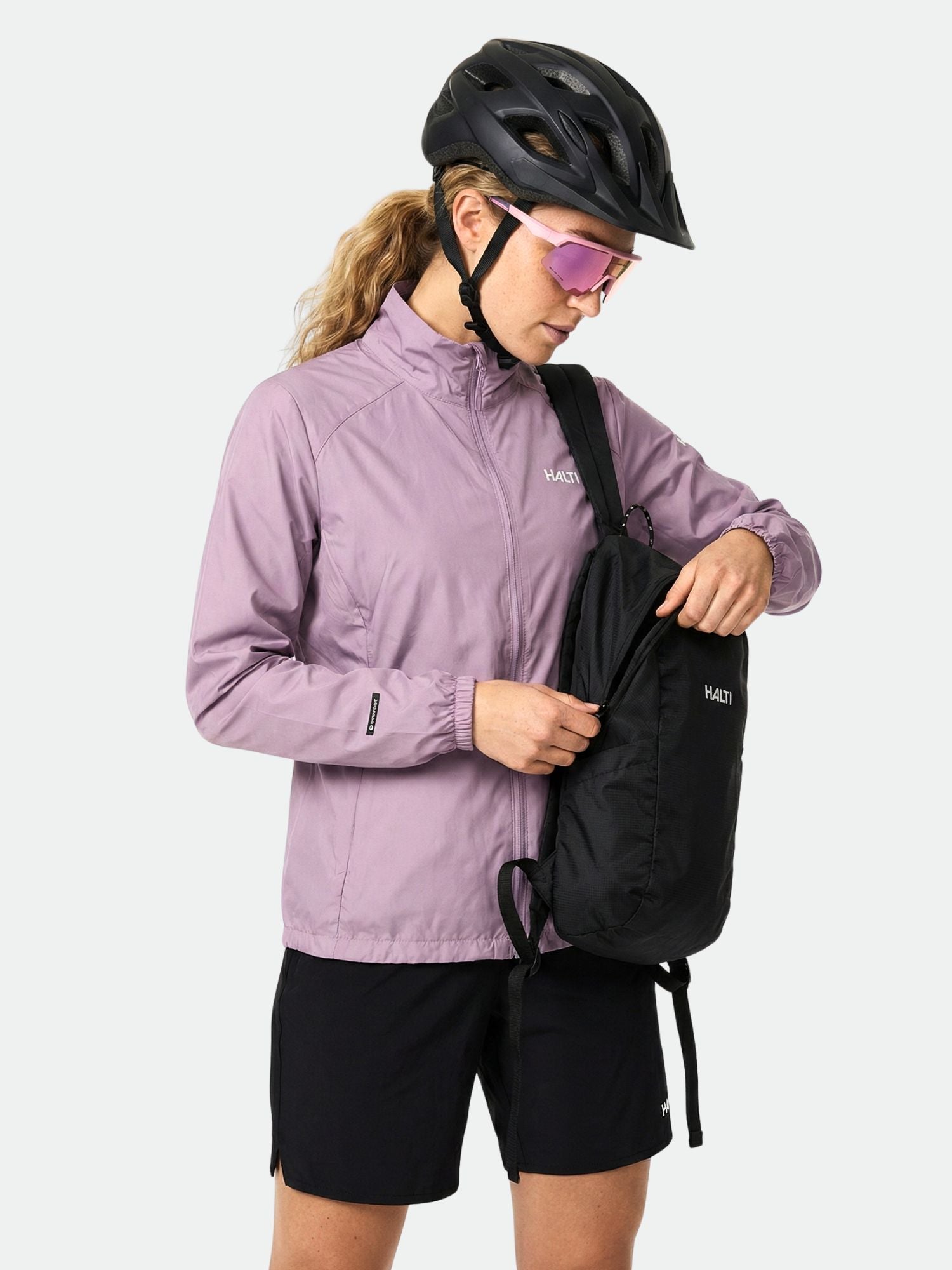 Eine Frau in einer Halti Saime Windbreaker Jacke Damen und schwarzen Shorts packt Gegenstände in einen schwarzen Rucksack.