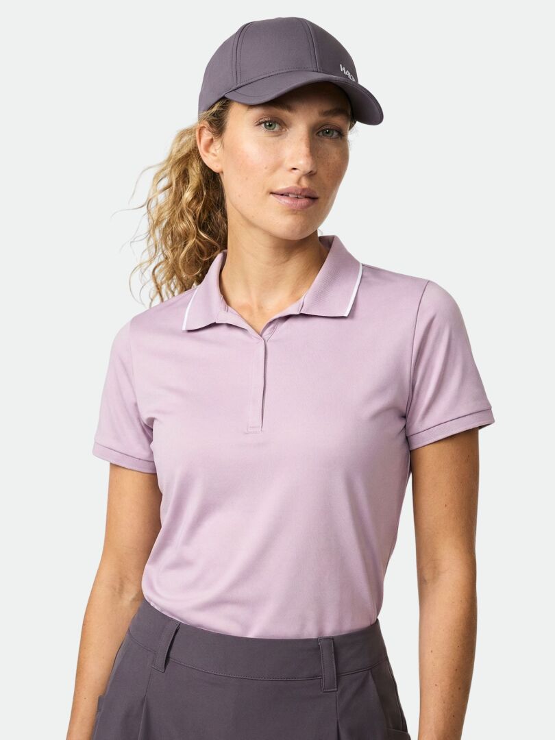 Eine Frau in einem hellvioletten Halti Swingi Technisches Poloshirt Damen und violetter Mütze steht vor einem schlichten Hintergrund.