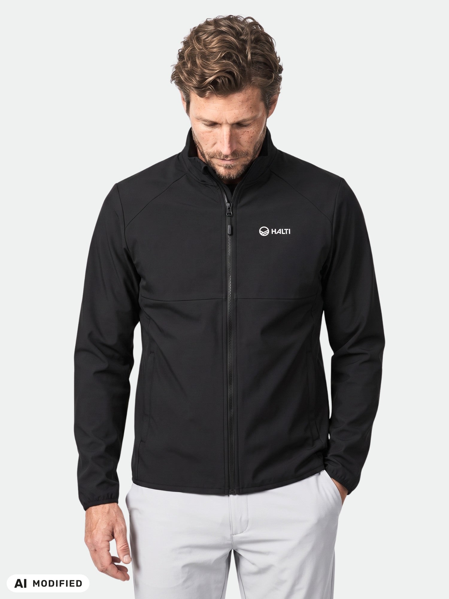 Ein Mann, der eine schwarze „Halti Wedge Softshell Jacke Herren“ und eine helle Hose trägt, steht vor einem schlichten weißen Hintergrund.