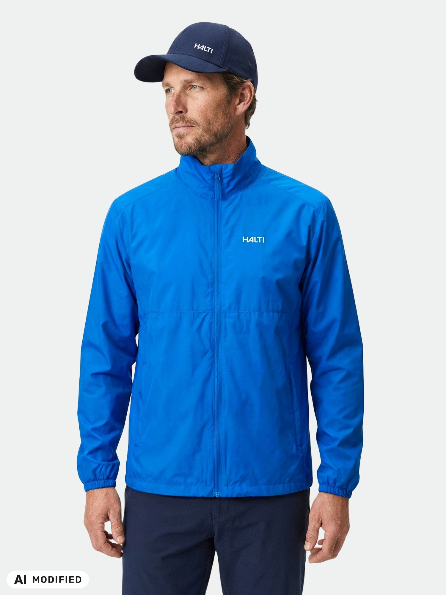 Ein Mann in der Halti Saime Windbreakerjacke für Herren und einer Mütze posiert vor einem weißen Hintergrund für Outdoor-Aktivitäten.