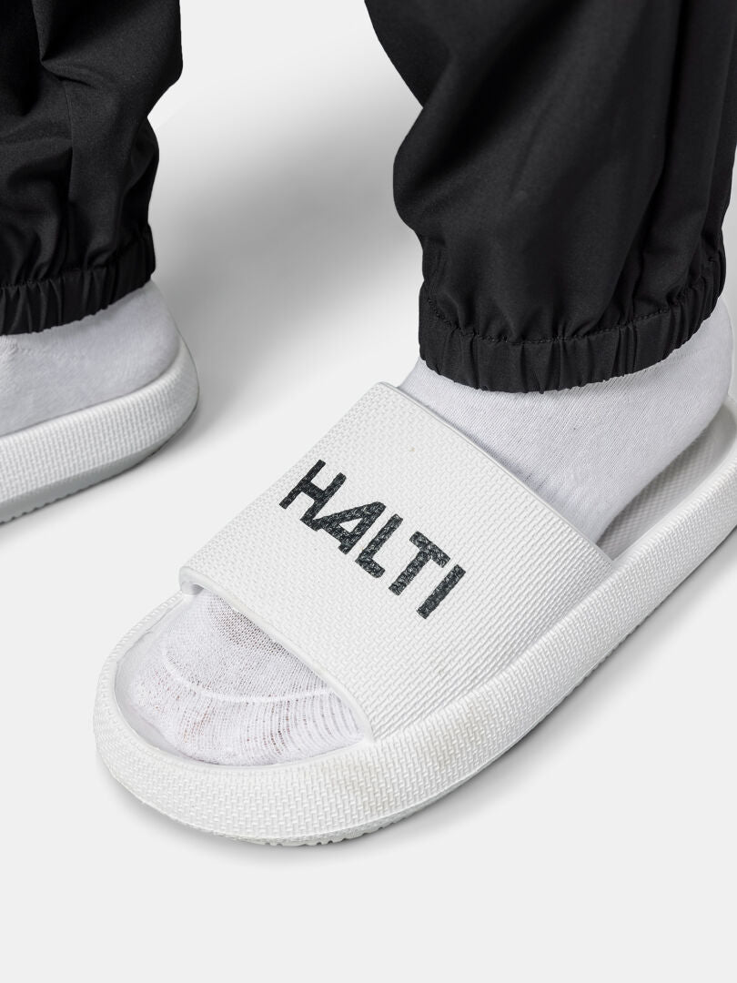 Nahaufnahme der Füße in weißen Socken und Halti-Slides, gepaart mit Halti Grets Jogginghose Damen in Schwarz, die an den Knöcheln gerafft ist.